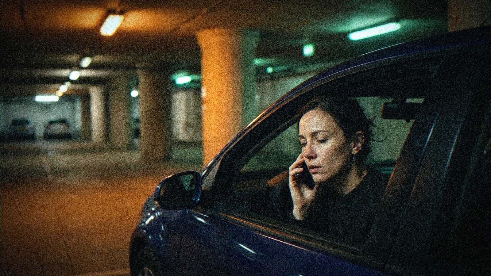 Créatrice seule assise dans sa voiture garée dans un parking souterrain la nuit, téléphone contre l'oreille pour vérifier un mix son sur un petit haut-parleur