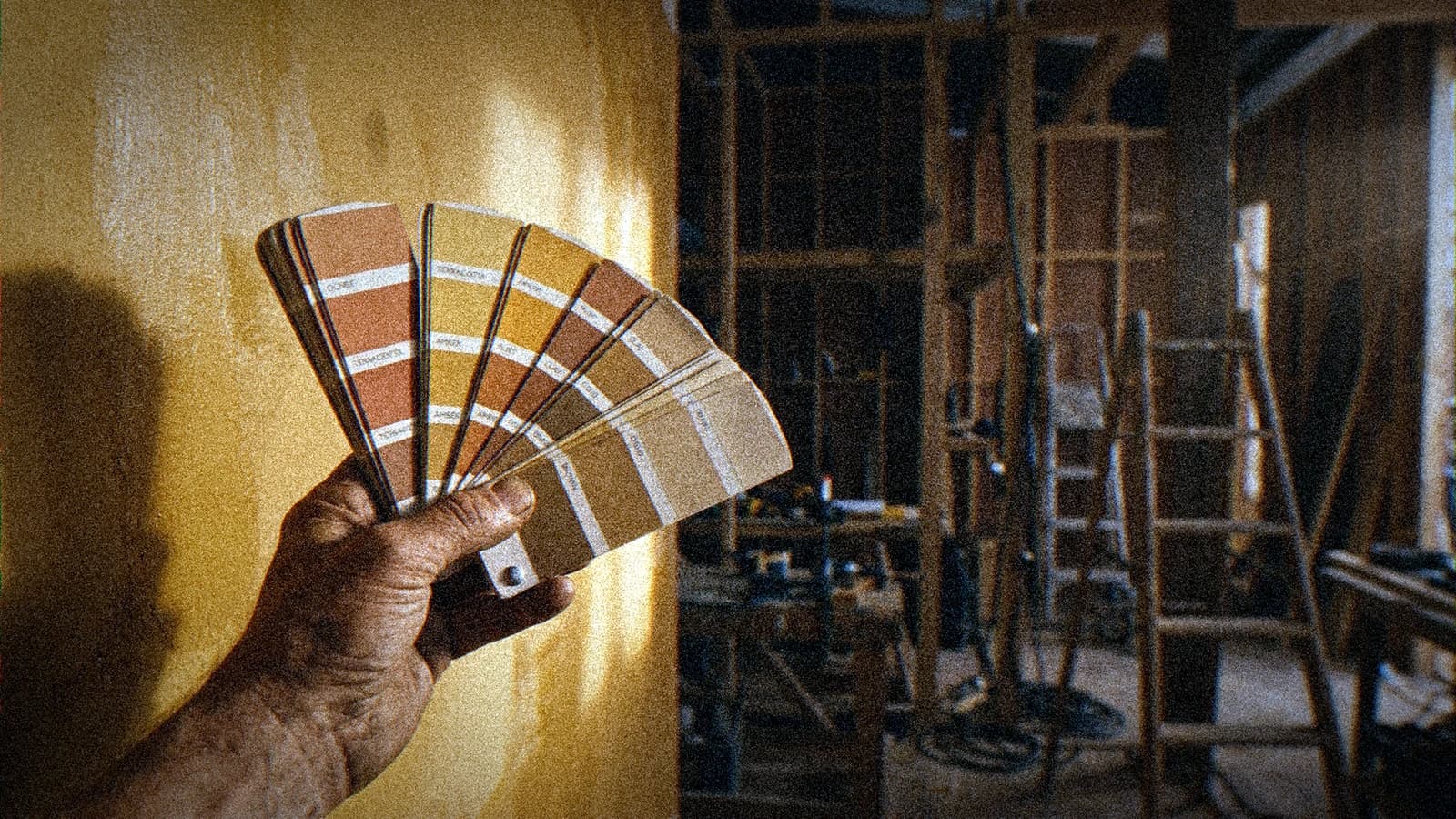 Main tenant un nuancier de couleurs déployé devant un mur fraîchement peint sur un décor en construction, lumière chaude de fin d'après midi